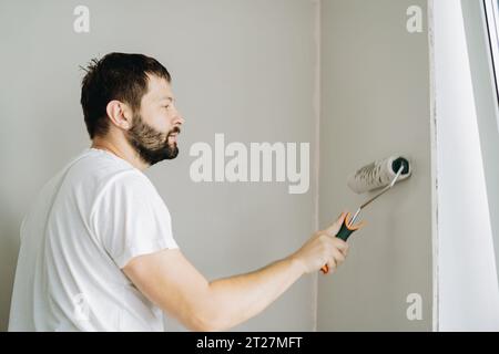 homme barbu caucasien peignant mur avec rouleau à peinture. Peinture appartement, rénovation de la maison avec de la peinture de couleur grise. Image avec mise au point sélective Banque D'Images