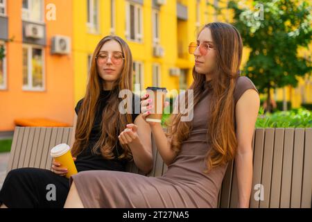 Mignonnes jeunes jumelles Gen Z avec des lunettes de soleil assis sur un banc et parlant sur la tenue de tasses à café jetables en papier dans le parc de la ville à l'extérieur Banque D'Images