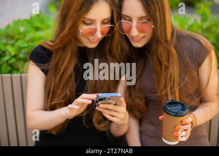 Des soeurs jumelles émerveillées avec des lunettes de soleil en utilisant les médias sociaux avec un téléphone intelligent assis sur un banc en ville une tasse à café en papier jetable tenant à l'extérieur Banque D'Images