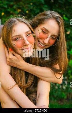 Stupéfié jeunes sœurs jumelles heureuses génération Zsnuggled avec des lunettes de soleil dans un parc, les femmes posant à l'extérieur. Concept d'amitié Happy Girls embrassant Banque D'Images