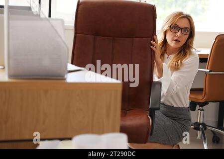 Femme d'affaires mature effrayée se cachant derrière la chaise dans le bureau Banque D'Images