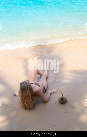 Une femme en maillot de bain se couche sur le sable à l'ombre d'un palmier et regarde la mer. Vacances dans les pays tropicaux. Banque D'Images