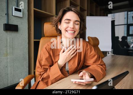 Portrait de femme riant sur la réunion, utilisant smartphone et écouteurs sans fil pour parler avec partenaire d'affaires, assis dans le bureau portant décontracté Banque D'Images