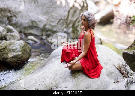 Femme religieuse faisant du yoga en longue robe rouge, dans la nature sauvage, près de la cascade en été. Banque D'Images
