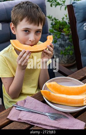 Angle élevé de mignon garçon préadolescent regardant la caméra assis à table et mangeant des fruits de melon de musc frais tranchés sur une plaque avec des sphères de melon dans le b glacé Banque D'Images