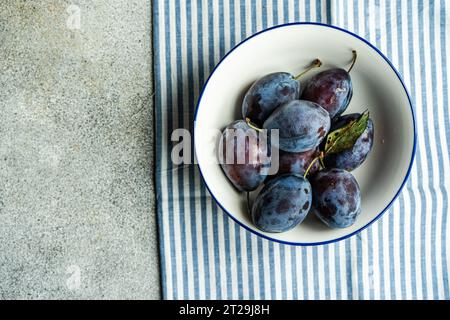 Vue de dessus des prunes mûres servies dans un bol en céramique sur une serviette rayée contre une surface grise Banque D'Images
