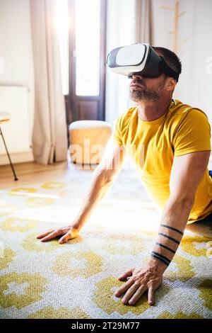 FIT gars barbu dans T-shirt jaune dans des lunettes VR faisant Cobra pose sur le tapis dans la pièce confortable lumineuse Banque D'Images