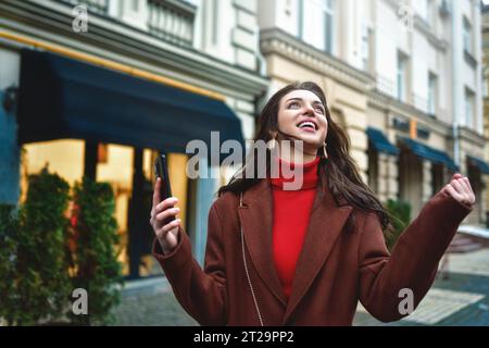 Femme branchée excitée célébrant de bonnes nouvelles tenant le téléphone dans la rue du soir Banque D'Images