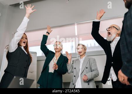 Vue à angle bas des employés enthousiastes multiethniques enthousiastes heureux levant les mains debout autour de la table de bureau dans une réunion d'affaires d'entreprise. Banque D'Images