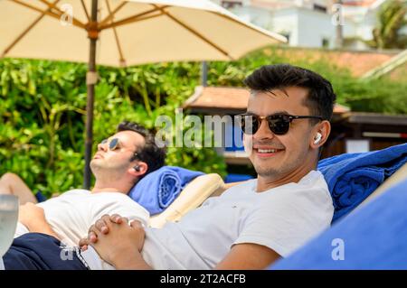 Un jeune homme d'une vingtaine d'années s'allonge sur un transat en écoutant de la musique avec AirPods avec son frère en arrière-plan dans un complexe hôtelier à Mui ne, au Vietnam. Banque D'Images