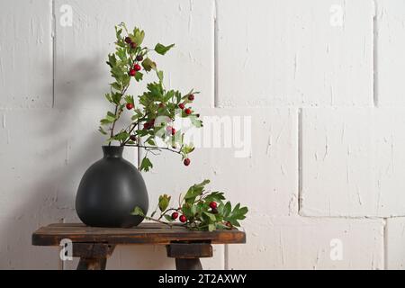 Branches d'aubépine avec des fruits rouges dans un vase noir sur un tabouret en bois rustique contre un mur blanc rugueux, décoration d'automne et d'hiver pour Noël Banque D'Images