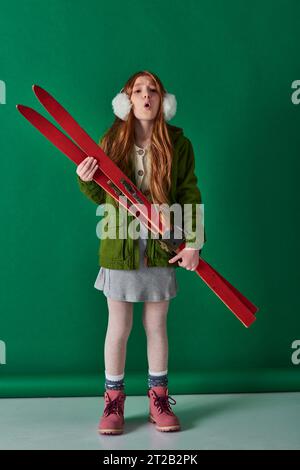 air froid, fille preteen dans les cache-oreilles et tenue d'hiver respirant et tenant des skis rouges sur turquoise Banque D'Images