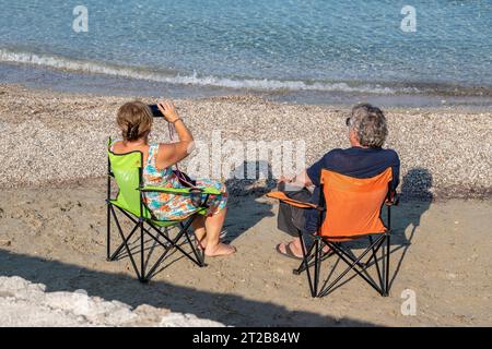 couple plus âgé ou d'âge moyen assis sur une plage dans des chaises pliantes aux couleurs vives regardant à travers des jumelles en vacances sur zakynthos grèce. Banque D'Images