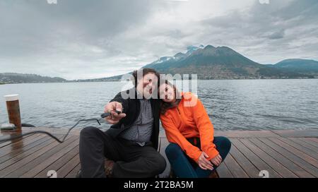 Jeune couple latino-américain assis sur une jetée en bois sur le lac San Pablo prenant un selfie avec le volcan Imbabura en arrière-plan par une journée nuageuse Banque D'Images