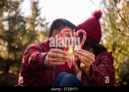 La canne à bonbons blanc rouge est tenue dans les mains tendues d'un couple amoureux dans des chemises à carreaux, des chapeaux tricotés près du marché vert des arbres de Noël. Homme heureux Banque D'Images