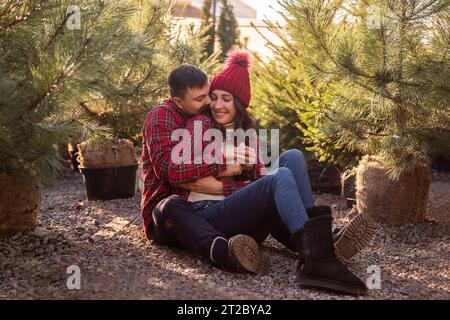 Portraits rapprochés de jeunes couples en chemises à carreaux rouges assis parmi les semis d'arbres de Noël au marché de rue. Homme embrasser la femme dans le chapeau tricoté W Banque D'Images