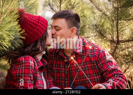 Joyeux jeune couple dans des chemises à carreaux rouges s'embrassant, tenant des étincelles festives dans les mains dans la forêt. Homme et femme rient au marché des arbres de Noël. CLO Banque D'Images