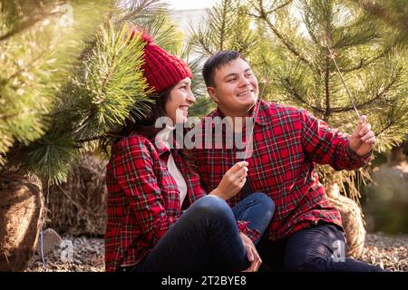 Joyeux jeune couple en chemises à carreaux rouges tient les étincelles festives en mains dans la forêt. Homme et femme rient au marché des arbres de Noël. Gros plan portra Banque D'Images