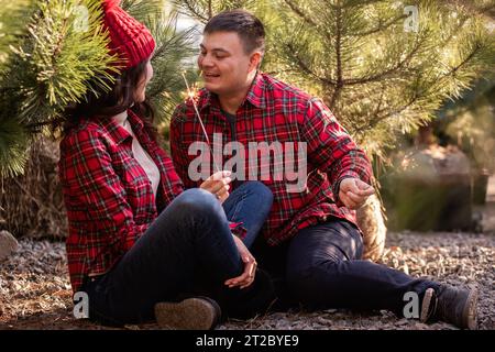 Joyeux jeune couple en chemises à carreaux rouges tient les étincelles festives en mains dans la forêt. Homme et femme rient au marché des arbres de Noël. Gros plan portra Banque D'Images