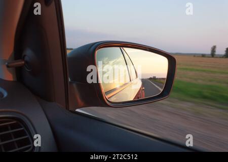 Photo d'une voiture prise à travers le rétroviseur latéral, coucher de soleil et ciel reflété dans le rétroviseur de la voiture. Banque D'Images