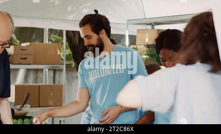 Homme caucasien âgé sur des béquilles est assisté par une femme noire amicale à une collecte de nourriture caritative. Jeunes volontaires qui apportent une aide humanitaire aux nécessiteux, aux sans-abri et aux handicapés. Prise de vue sur trépied. Banque D'Images