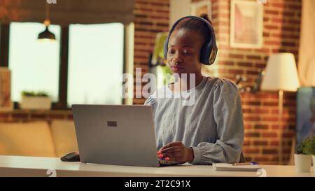 Télétravailleur écoutant de la musique et résolvant des tâches quotidiennes, femme indépendante. Fille afro-américaine s'amusant avec casque pendant qu'elle travaille sur des tâches pendant l'heure de coucher du soleil à la maison. Prise de vue à main levée. Banque D'Images
