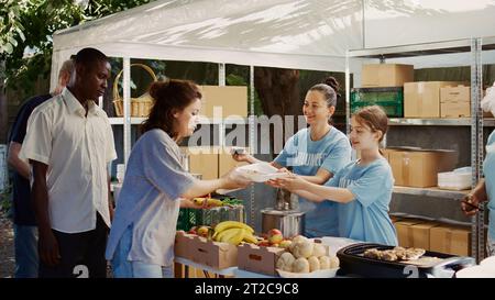 Une jeune fille caucasienne emballe gratuitement de la nourriture à distribuer aux défavorisés avec l'aide d'autres travailleurs caritatifs. Des femmes bénévoles servant des repas chauds aux sans-abri et affamés à la banque alimentaire extérieure. Banque D'Images