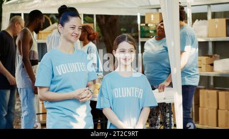 Portrait en vue latérale d'une mère et de sa fille en soutien à une campagne extérieure de banque alimentaire pour lutter contre la faim. Deux femmes caucasiennes portant des t-shirts bénévoles bleus sont prêtes à aider les sans-abri. Banque D'Images