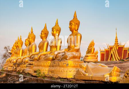 Rangées de belles, miniatures, sculptures d'or vibrantes de Bouddha, illuminées par le soleil couchant, alignées au sommet d'un mur en pente, à côté du temple, Banque D'Images