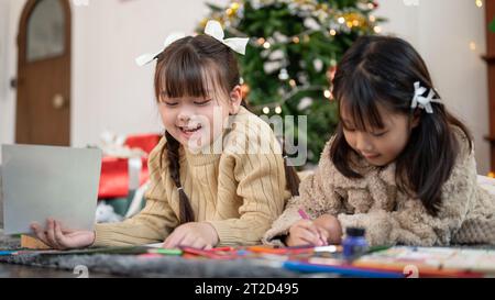 Deux adorables et joyeuses jeunes filles asiatiques s'amusent, aiment parler et faire des cartes de Noël tout en étant allongées sur le sol du salon ensemble Banque D'Images