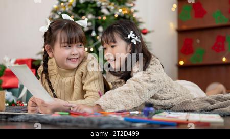 Deux adorables et joyeuses jeunes filles asiatiques s'amusent, aiment parler et faire des cartes de Noël tout en étant allongées sur le sol du salon ensemble Banque D'Images