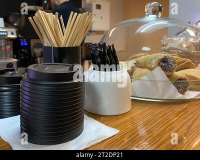 Bâtons jetables en bois naturel pour mélanger le sucre dans le thé ou le café, sucrier, couvercles thermo en plastique pour tasses et gâteaux sur le bar dans un petit café. Banque D'Images