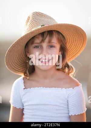 Souriant jolie fille de 8-9 ans dans un chapeau de paille et une robe blanche regarder la caméra. Saison estivale. Enfance Banque D'Images