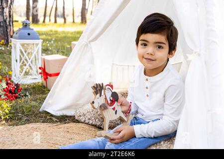 Garçon souriant d'enfant jouant avec la décoration de Noël dans la tente extérieure de tipi. Photoshoot familial hivernal dans la forêt. Banque D'Images