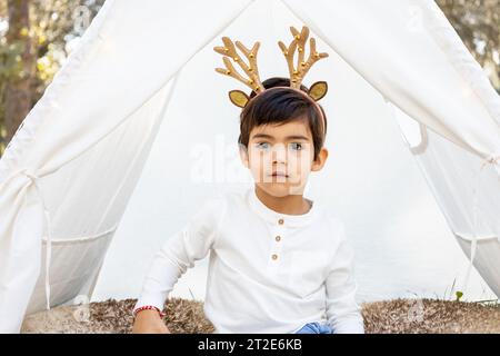 Enfant garçon souriant jouant avec la décoration de Noël dans la tente extérieure en tipi avec des bois de renne scintillants. Photoshoot familial hivernal dans la forêt. Banque D'Images