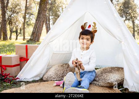 Enfant garçon souriant jouant avec la décoration de Noël dans la tente extérieure en tipi avec des bois de renne scintillants. Photoshoot familial hivernal dans la forêt. Banque D'Images