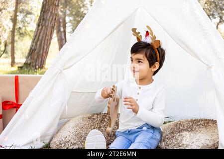 Enfant garçon souriant jouant avec la décoration de Noël dans la tente extérieure en tipi avec des bois de renne scintillants. Photoshoot familial hivernal dans la forêt. Banque D'Images