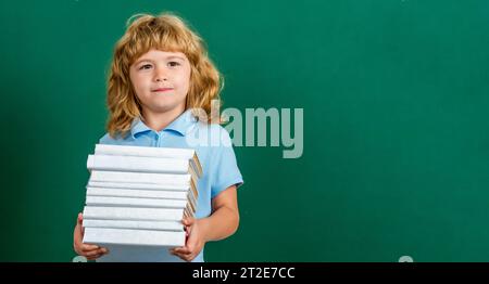 Écolier en classe. Enfant tenant la pile de livres avec le panneau de mortier sur le tableau noir. Retour à l'école. Enfant garçon contre tableau noir vert. Éducation et Banque D'Images