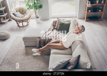 Photo vue de dessus de positive brillante dame âgée porter un cardigan blanc relaxant les yeux fermés bras derrière la tête à l'intérieur de la chambre d'appartement Banque D'Images