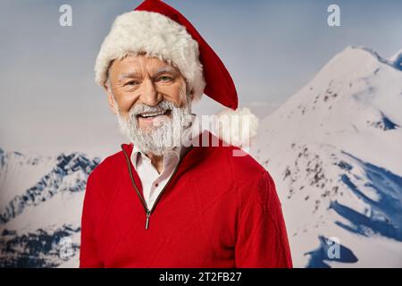 Beau père Noël joyeux en tenue rouge souriant à la caméra avec toile de fond de montagne, concept d'hiver Banque D'Images