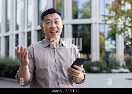 Portrait d'un jeune homme asiatique heureux employé de bureau debout près d'un centre de bureau et tenant un téléphone. Banque D'Images