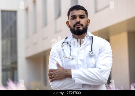 Jeune médecin musulman sérieux debout à l'extérieur de la clinique à l'extérieur portant une robe et tenant un stéthoscope et croisant ses bras regardant avec confiance la caméra. Banque D'Images