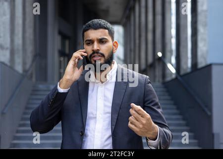 Bouleversé jeune homme d'affaires indien parlant au téléphone dans la rue près du bureau, salle d'audience. Insatisfait et gesticulant avec ses mains. Banque D'Images