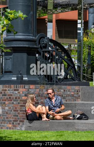 Un jeune couple se détendant au soleil près de la structure de support de gaz de fer réutilisée à Gasholder Park, Kings Cross, Londres. Banque D'Images