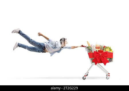 Plan en longueur d'un jeune homme occasionnel volant et tenant un chariot avec de la nourriture et un arc de ruban rouge isolé sur fond blanc Banque D'Images