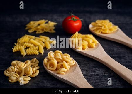 Augsbourg, Bavière, Allemagne. 19 octobre 2023. Trois cuillères en bois avec des nouilles crues. Pâtes fusilli, farfalle et trulli décorées sur des cuillères en bois sur une table sombre rustique avec une tomate rouge fraîche *** Drei Holzlöffel mit rohen Nudeln. Fusilli, Farfalle und Trulli Pasta dekoriert auf Holzlöffeln auf einem rustikalen dunklen Tisch mit einer frischen roten tomate crédit : Imago/Alamy Live News Banque D'Images