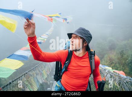 Jeune homme souriant traversant le canyon au-dessus du pont suspendu et touchant des drapeaux de prière tibétains multicolores articulés au-dessus de la gorge. Route d'escalade de pic de Mera tr Banque D'Images