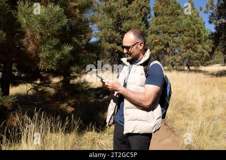 Sourire 40 yo Homme dans lunettes de soleil tenant smartphone, Gadget. Sapins, forêt sur fond. La nature en automne feuilles saison. Horizontal. Actif Banque D'Images