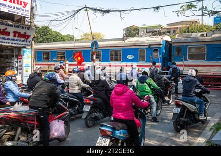 Un train des chemins de fer du Vietnam passe des gens à moto et à vélo à un passage à niveau (passage à niveau) à Hanoi, Vietnam. Banque D'Images