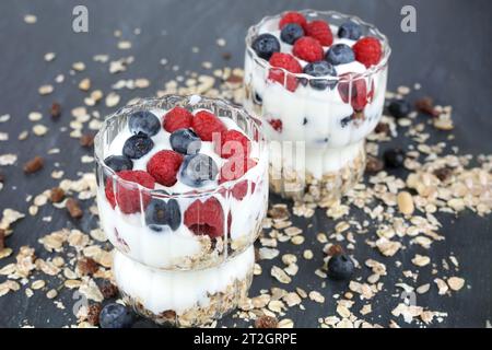 Muesly avec des framboises et des bleuets dans deux verres sur fond sombre. Banque D'Images